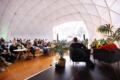 The inside of giant white domed tent with people sitting onstage and other people sitting at chairs in the audience watching the onstage speakers