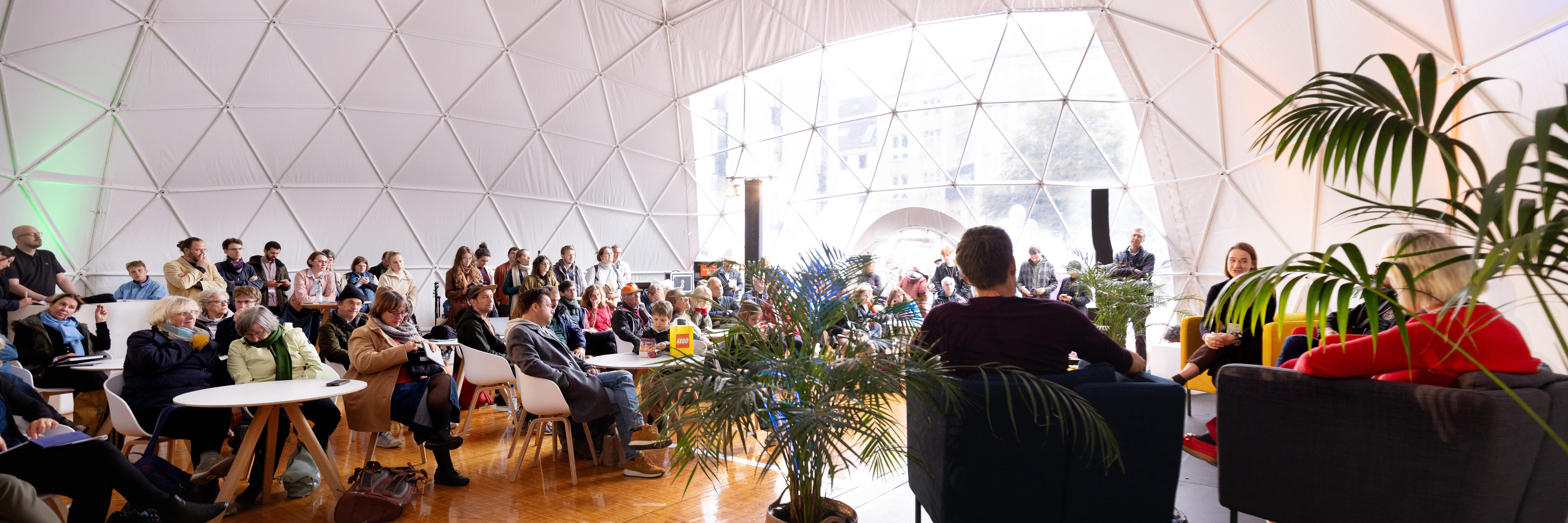 The inside of giant white domed tent with people sitting onstage and other people sitting at chairs in the audience watching the onstage speakers