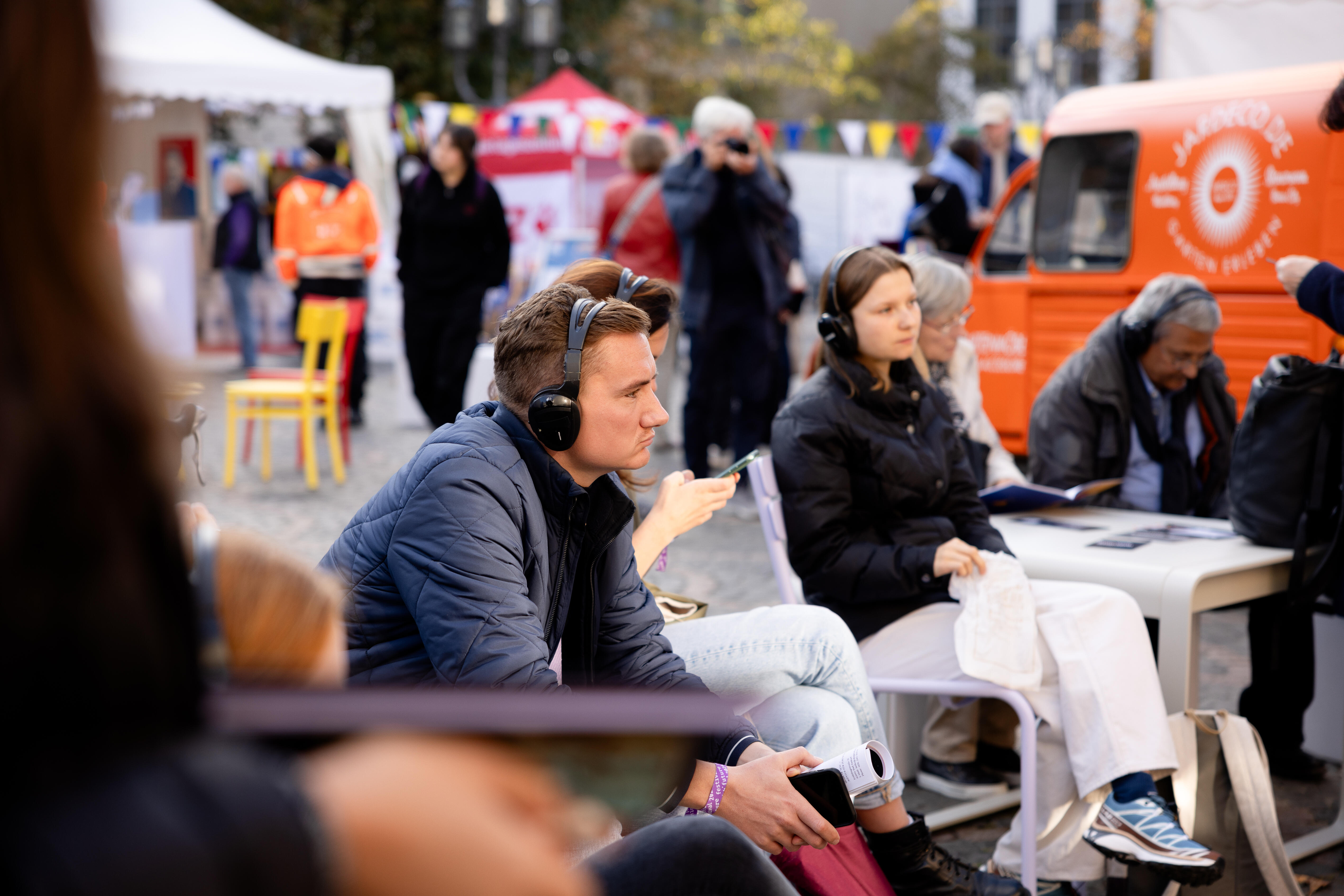 Ein Mann sitzt inmitten einer Gruppe von Menschen, die Kopfhörer tragen und aufmerksam etwas hören, während im Hintergrund Menschen auf einem Festival mit Zelten und einem Lastwagen herumlaufen