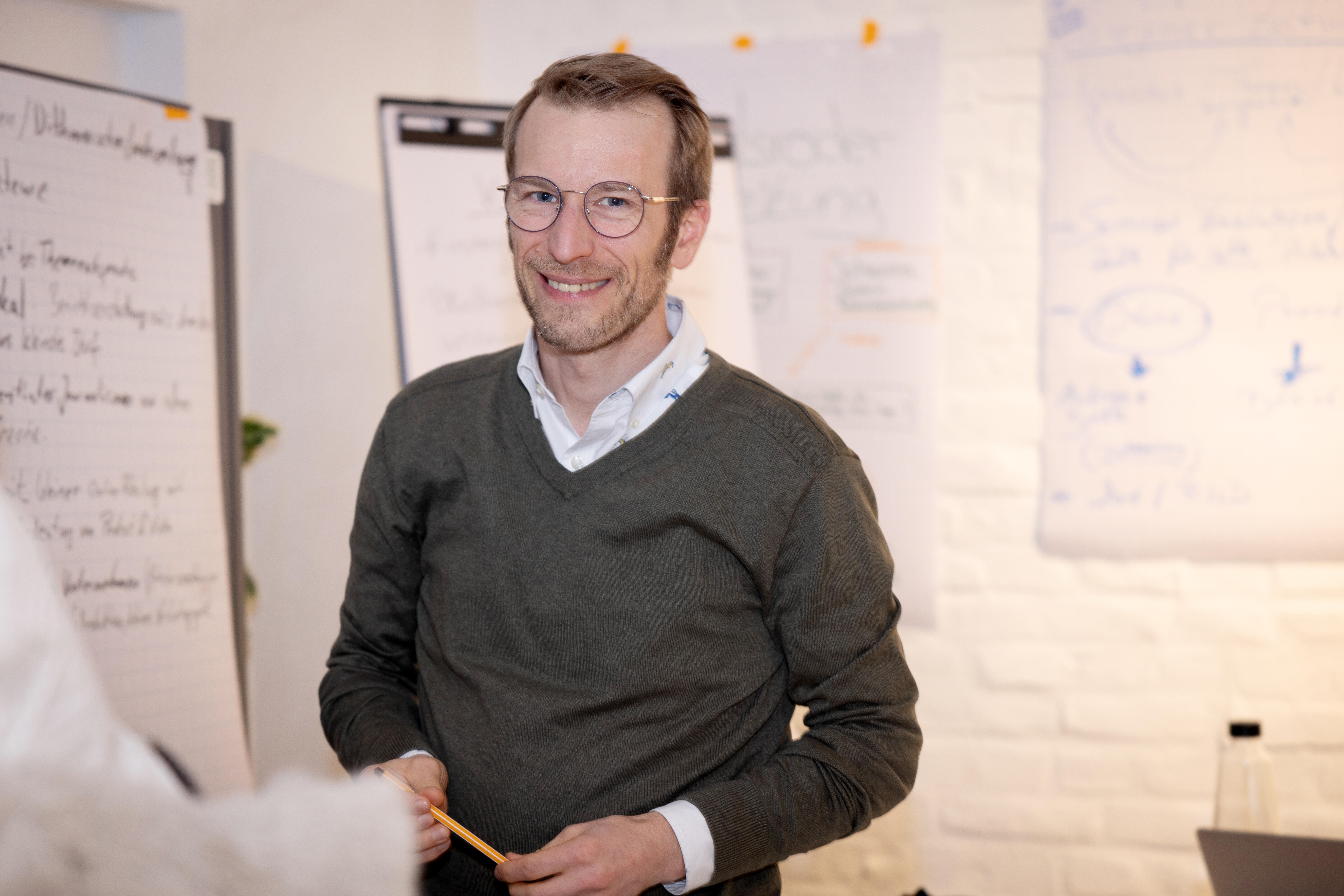 A man holds a pencil in front of flip charts and smiles